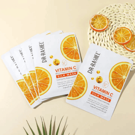 A hand holding multiple Dr. Rashel Vitamin C Brightening and Anti-Aging Silk Mask sachets against a soft yellow background, showing packaging with orange graphics and highlighting niacinamide for skin brightening and anti-aging benefits.