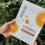 A hand holding a box of Dr. Rashel Vitamin C Brightening and Anti-Aging Silk Mask against a leafy outdoor background, featuring citrus-themed packaging and highlighting skincare benefits.
