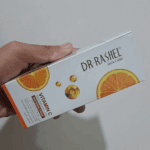 Hand holding a Dr. Rashel Vitamin C Brightening & Anti-Aging Whitening Cream box with orange slice graphics on clean white packaging.
