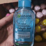 Hand holding Garnier Micellar Cleansing Water with Salicylic BHA surrounded by decorative items, removes makeup, cleanses and helps reduce acne and dark spots.
