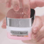 Hand holding Glycolic-Bright Glowing Cream Day SPF 17 moisturizer with a silver lid and pink jar.