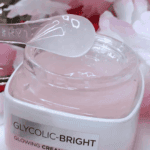 Close-up of Glycolic-Bright Glowing Cream Day SPF 17 with a spatula scooping pink gel cream from the jar, surrounded by soft pink flowers.