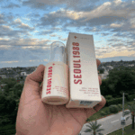 Hand holding K-Secret Seoul 1988 Retinal Liposome 2% + Black Ginseng Serum 30ml bottle with its box packaging outdoors with city view in the background.