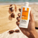 Hand holding La Roche-Posay Anthelios SPF 50 sunscreen bottle at the beach with sunglasses and sand blurred in the background.