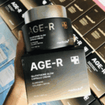 Hand holding a jar of Medicube AGE-R Glutathione Glow Capsule Cream placed on its box, with multiple AGE-R product boxes displayed in the background.