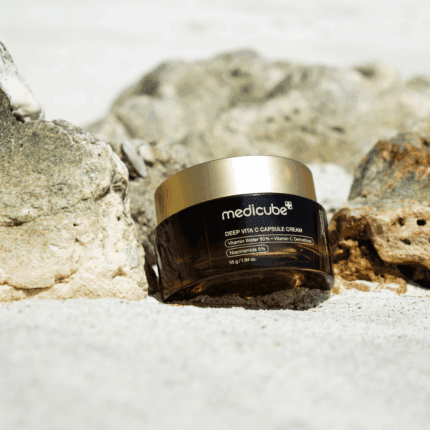 Medicube Deep Vita C Capsule Cream vitamin C moisturizer in a dark amber jar with a gold lid placed on sand between natural rocks.