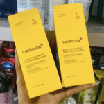Hand holding two yellow Medicube Kojic Acid Turmeric Night Wrapping Mask boxes (75 ml) in front of a shelf with skincare products, highlighting ingredients like kojic acid, vitamin C, retinol, turmeric, and collagen for overnight skin care.