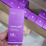Hand holding Medicube Retinol NMN Boosting Serum 1.9% box with multiple purple product boxes in a shipping carton in the background.