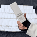 A hand holding a box of The Ordinary Hyaluronic Acid 2% + B5 serum, with multiple identical product boxes arranged in the background, highlighting the minimalist white packaging and hydration-focused skincare formula.