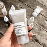 Hand holding The Ordinary Natural Moisturizing Factors + HA 30ml face moisturizer with skincare products in background