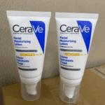 Two CeraVe Facial Moisturising Lotion AM SPF 50 pump tubes placed side by side on a surface against a neutral textured background, showing packaging for normal to dry skin with UV protection.